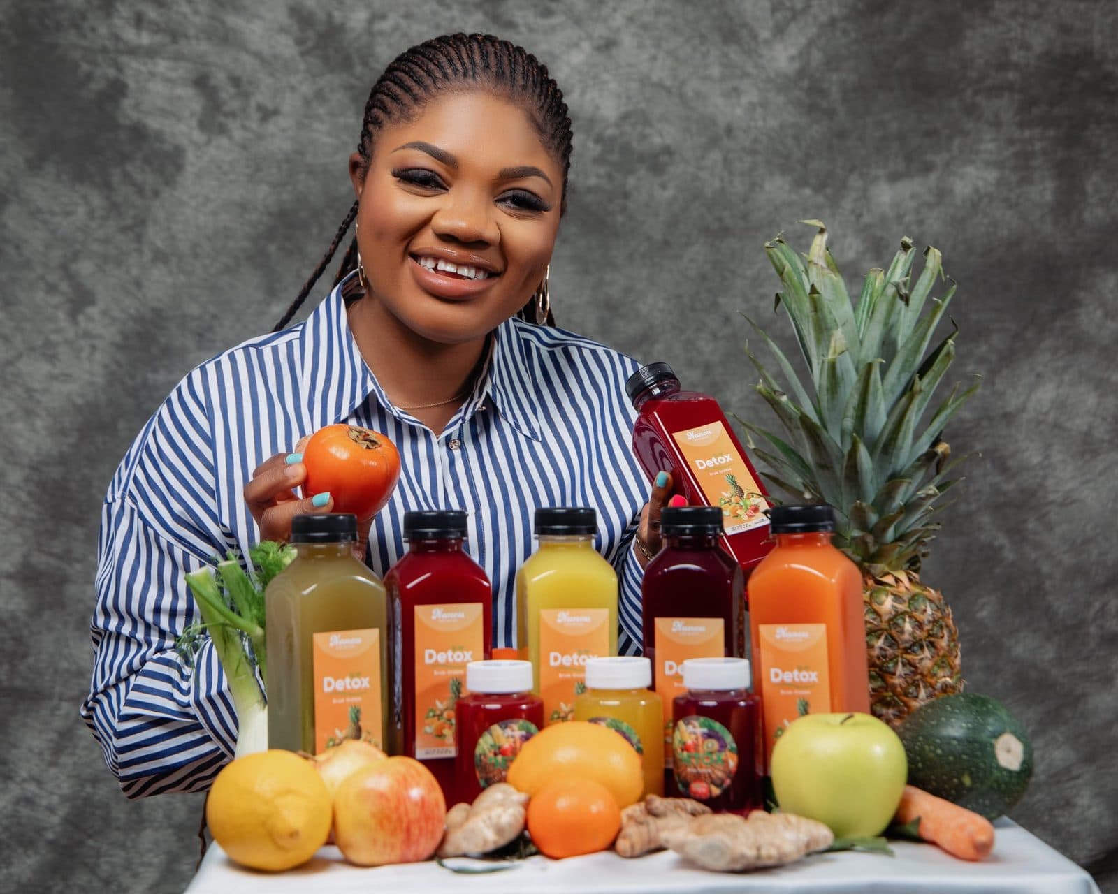 La fondatrice de Nanou Detox derrière cinq bouteilles de jus, entourées d'ananas, d'agrumes, de gingembre, de pommes et de carottes.